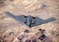 A B-2 Spirit stealth bomber flying with GBU-57 MOP explosions detonating across a desert landscape below.