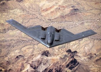 A B-2 Spirit stealth bomber flying with GBU-57 MOP explosions detonating across a desert landscape below.