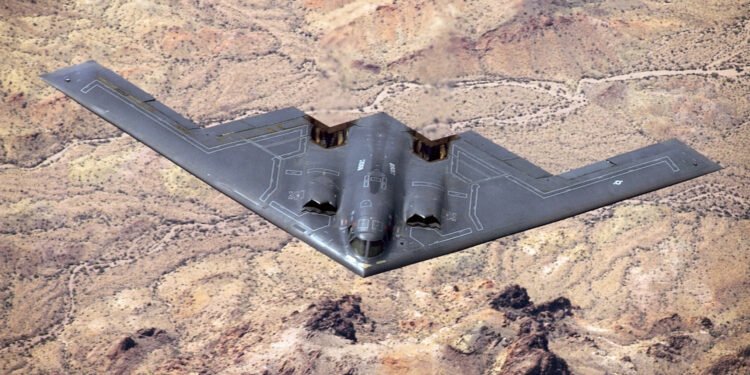 A B-2 Spirit stealth bomber flying with GBU-57 MOP explosions detonating across a desert landscape below.