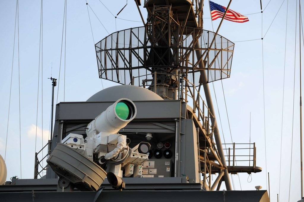 A white, tubular directed energy weapon turret mounted on the deck of a naval warship against a clear sky.