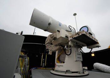 A white, tubular directed energy weapon turret mounted on the deck of a naval warship against a clear sky.