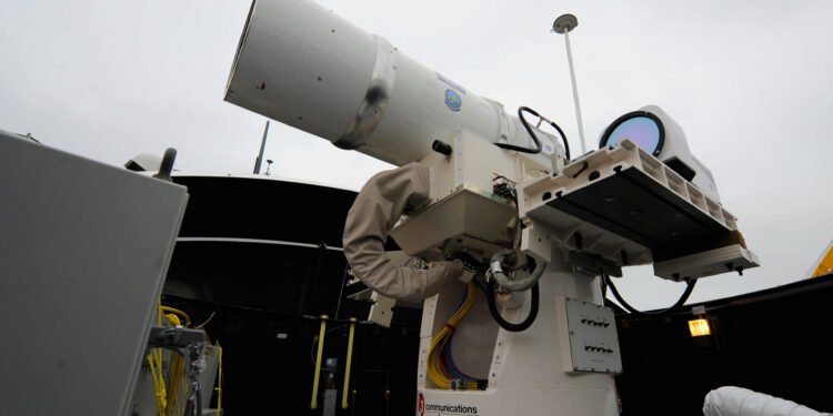 A white, tubular directed energy weapon turret mounted on the deck of a naval warship against a clear sky.