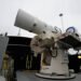 A white, tubular directed energy weapon turret mounted on the deck of a naval warship against a clear sky.