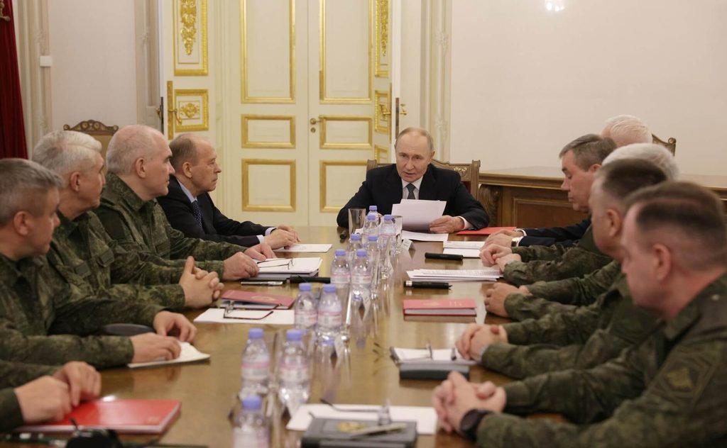 Russian President Vladimir Putin sitting at a long table during a defense briefing with military leadership.